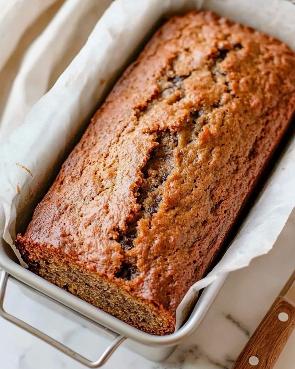 A freshly baked loaf of banana bread with a golden brown, slightly cracked top showing a moist, dense texture inside, sitting in a white metal loaf pan lined with parchment paper. The bread has small dark nut pieces visible throughout the top layer, adding a bit of texture. The loaf pan is placed on a white marbled surface next to a wooden-handled knife with a silver blade. The image captures a close-up view highlighting the rough, crumbly top crust and the soft interior. Photo taken with an iphone --ar 4:5 --v 6.1