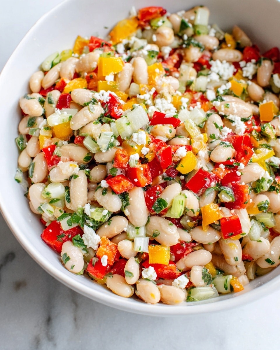 A close-up of a colorful bean salad in a white bowl, showing a mix of three layers: a base of light beige beans, topped with bright small pieces of red and yellow bell peppers, white onion, celery, and chopped green herbs scattered evenly throughout. Tiny white crumbles are sprinkled on top, adding a soft texture to the salad. The bowl sits on a white marbled surface. photo taken with an iphone --ar 4:5 --v 7