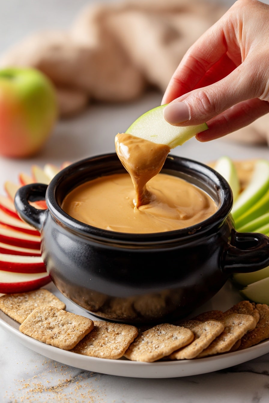 A small black pot with a handle holds a thick, creamy, light brown dip with a smooth texture, and a woman's hand is dipping a green apple slice into it. The pot is placed on a white plate with dry beige crackers in the foreground, red apple slices to the left, and more green apple slices to the right. The background shows a soft, warm light and a white marbled surface. Photo taken with an iphone --ar 2:3 --v 7 - Creamy Pumpkin Dip with Spices and Marshmallow Cream, pumpkin dip with spices and marshmallow fluff, autumn pumpkin dessert dip, cozy pumpkin appetizer, easy pumpkin party dip