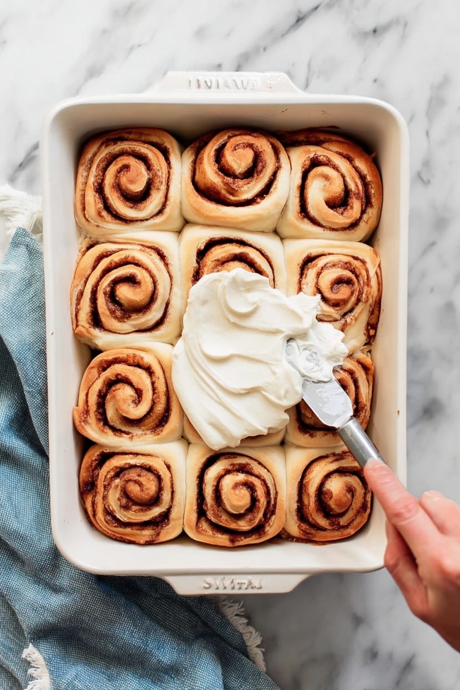 A white rectangular baking dish holds twelve cinnamon rolls arranged in a 3 by 4 grid. Each roll shows a spiral pattern with light brown dough and darker brown cinnamon filling. A woman's hand is spreading smooth white frosting over the center rolls with a silver spreading knife. The dish sits on a white marbled surface with a folded blue cloth nearby. Photo taken with an iphone --ar 2:3 --v 7 - Homemade Cinnamon Rolls with Glaze, cinnamon rolls recipe, soft cinnamon rolls, easy cinnamon roll recipe, homemade pastry treats