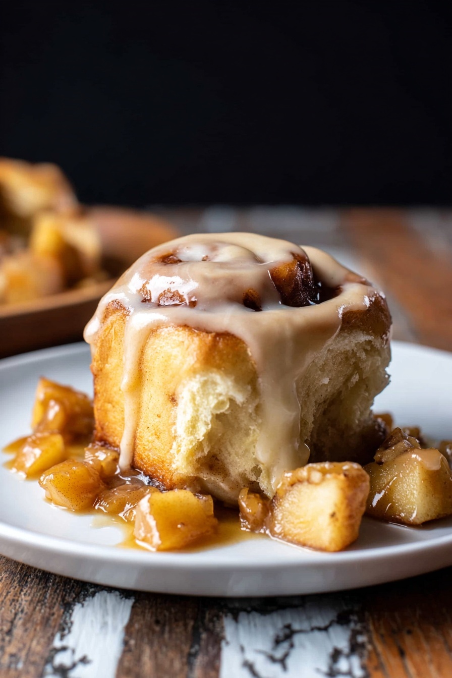 A cinnamon roll with visible swirls and a light golden brown color, topped with thick creamy icing that drapes over the edges, sitting on a white plate. Surrounding the roll are several torn pieces showing a soft, fluffy inside with a slightly darker cinnamon layer. Small chunks of a caramel-like sauce with bits of apple sit around the base, adding a shiny texture. The background is dark, and the plate rests on a rustic wooden surface with a white marbled texture. photo taken with an iphone --ar 2:3 --v 7 - Apple Cinnamon Rolls with Brown Butter Maple Icing, fall-inspired cinnamon rolls, homemade apple cinnamon pastry, brown butter maple icing recipe, cozy weekend breakfast