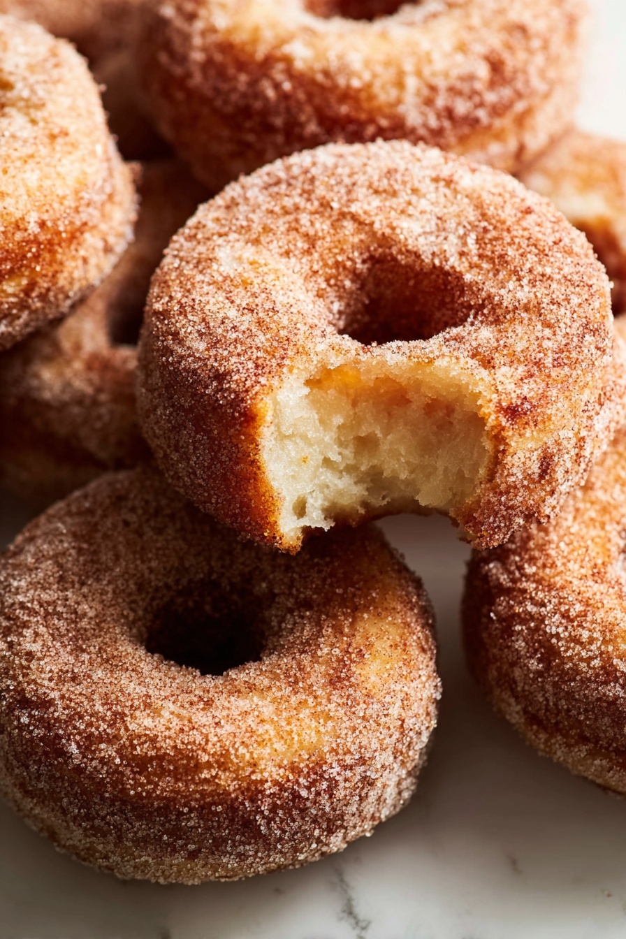 A stack of three sugar-covered donuts sits on a white marbled surface, with the top donut showing a bite taken out, revealing a soft, light brown inside. Each donut has a rough texture coated evenly in sugar, giving a slightly grainy look. The donuts are arranged one on top of the other, with the bitten one on the top, the middle and bottom donuts fully intact, all round with a visible center hole. The lighting softly highlights the sugar crystals and the golden brown color of the fried dough. photo taken with an iphone --ar 2:3 --v 7 - Apple Cider Donuts, homemade apple cider donuts, fall donut recipes, cinnamon apple donuts, soft fluffy apple cider treats