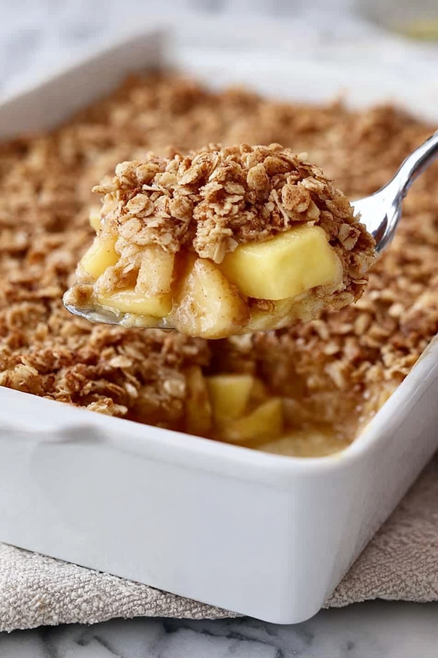 A white rectangular baking dish filled with a dessert that has three visible layers: at the bottom, soft light yellow chunks of apple, in the middle, a slightly sticky cinnamon-spiced syrup, and on top, a thick layer of golden brown oat crumble with a rough texture. A silver spoon is lifting a portion from the dish, showing the layers clearly. The dish is placed on a white marbled surface with a bit of a folded cloth underneath. Photo taken with an iphone --ar 2:3 --v 7 - Amish-Style Apple and Cinnamon Baked Oatmeal, baked oatmeal with apple and cinnamon, cozy healthy breakfast recipes, easy breakfast baking ideas, wholesome oat bake for mornings