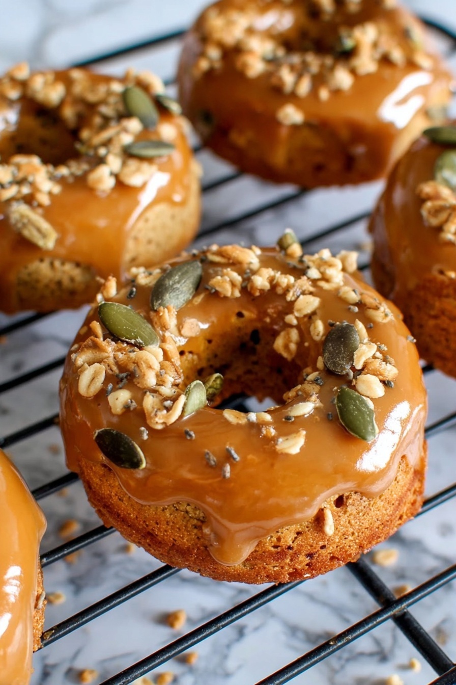 The image shows several small round donuts with a shiny caramel glaze on the top layer, sitting on a black cooling rack. The caramel glaze is smooth and glossy, covering the top and sides of the donuts. On top of the caramel layer, there are scattered crunchy toppings made of light brown crumbles and green pumpkin seeds, adding texture and color contrast. The donuts have a golden brown base with a soft, slightly rough surface. The background has a white marbled texture that frames the cooling rack and donuts softly. photo taken with an iphone --ar 2:3 --v 7 - Pumpkin Donuts with Bourbon Caramel Glaze, fall pumpkin donuts, bourbon caramel glaze recipe, cozy autumn desserts, easy pumpkin donut recipe