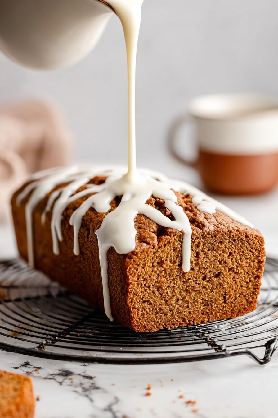 A loaf of brown textured bread sits on a round metal cooling rack with open grids. White icing is being poured over the top from above, flowing down the sides in thick, smooth ribbons. The loaf has a rough crust with deep cracks and a soft, porous interior visible on the side. In the background to the right, there is a blurred white cup with a brown lower half, all on a white marbled surface. Photo taken with an iphone --ar 2:3 --v 7 - Healthy Pumpkin Bread with Maple Glaze, pumpkin bread recipes, healthy fall desserts, wholesome pumpkin loaf, maple glaze pumpkin bread