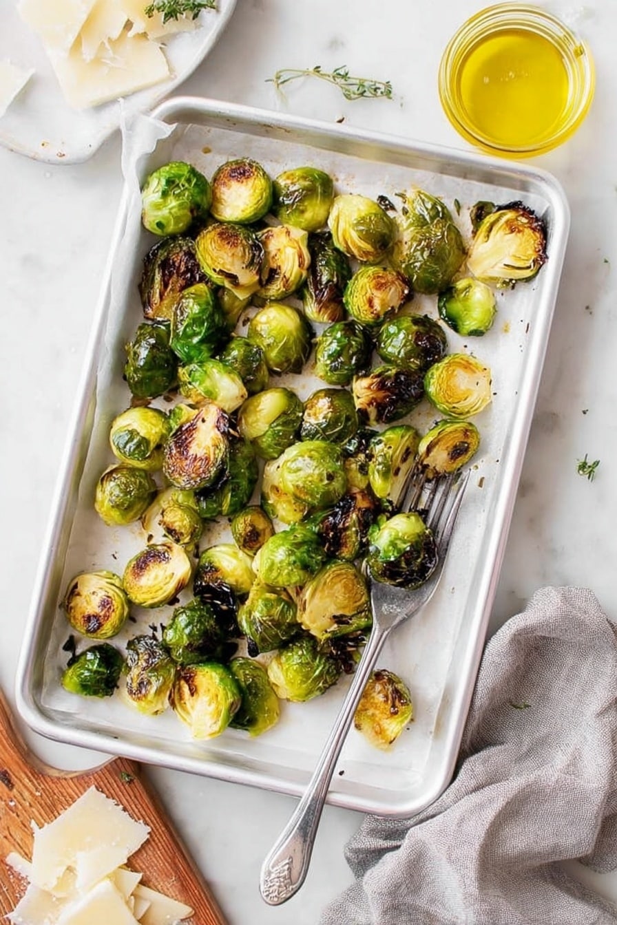 A white baking tray lined with white parchment paper holds a single layer of roasted Brussels sprouts, some halved and some whole, showing a mix of bright green and dark brown charred spots, giving a crispy texture. The tray is placed on a white marbled surface with a silver fork resting in the bottom right corner inside the tray. To the right, there is a small glass jar filled with yellow oil, and at the bottom left corner, part of a wooden board with thinly shaved white cheese is visible, along with a grey cloth nearby. The overall scene is bright and fresh, highlighting the crispy roasted Brussels sprouts. photo taken with an iphone --ar 2:3 --v 7 - Lemon Parmesan Roasted Brussels Sprouts, roasted Brussels sprouts with lemon and parmesan, healthy side dishes with brussels sprouts, lemon parmesan vegetable recipes, easy roasted Brussels sprouts