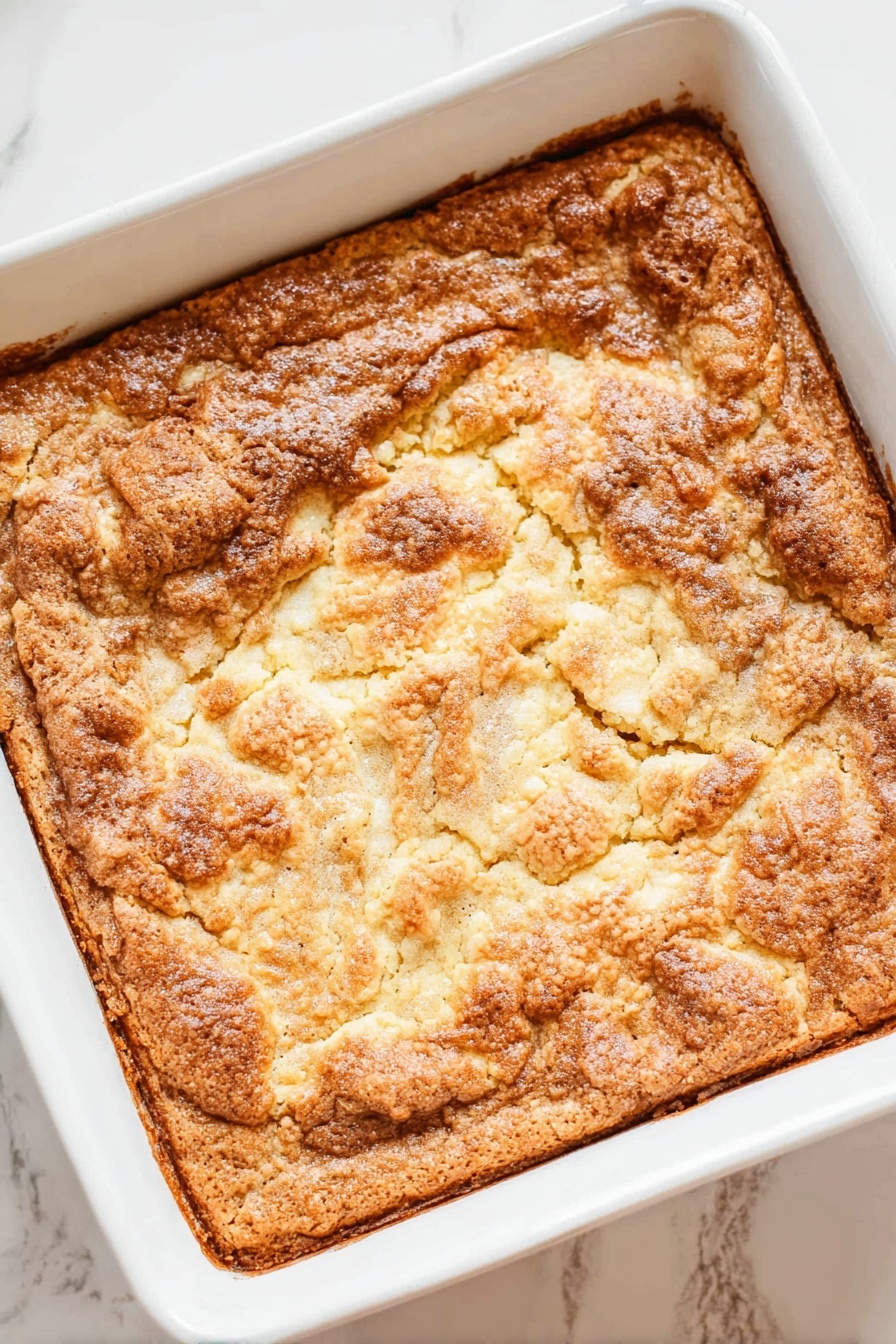 A square white baking dish holds a baked dessert with one thick layer that has a rough, cracked, golden-brown surface. The top looks crunchy and uneven with some lighter and darker brown spots, showing a slightly bubbled texture. The edges are more browned and crisp, while the center parts have a mix of smooth and bumpy patches. The dish is placed on a white marbled surface. Photo taken with an iphone --ar 2:3 --v 7 - Apple Dump Cake, easy apple dump cake, quick apple dessert, effortless apple cobbler, simple apple cake