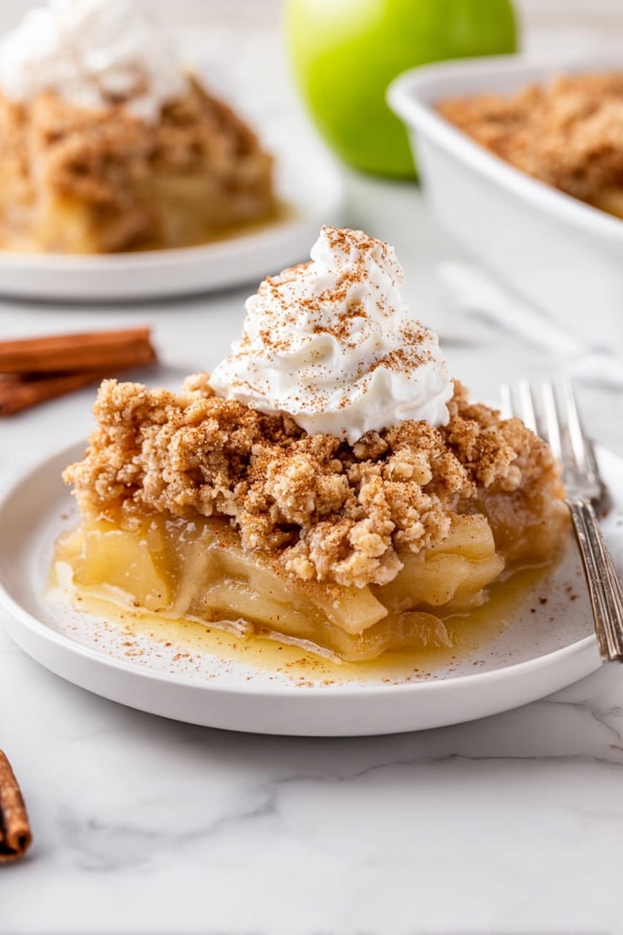 A slice of apple crumble sits on a white plate with three layers visible: a bottom layer of soft, cooked apple slices in a light golden sauce, a thick middle layer of crumbly, light brown streusel topping with a rough texture, and a dollop of white whipped cream sprinkled lightly with cinnamon on top. In the background, there is a second plate with more apple crumble and a silver fork. The scene is set on a white marbled surface with a halved green apple and a cinnamon stick placed nearby. Photo taken with an iphone --ar 2:3 --v 7 - Apple Dump Cake, easy apple dump cake, quick apple dessert, effortless apple cobbler, simple apple cake
