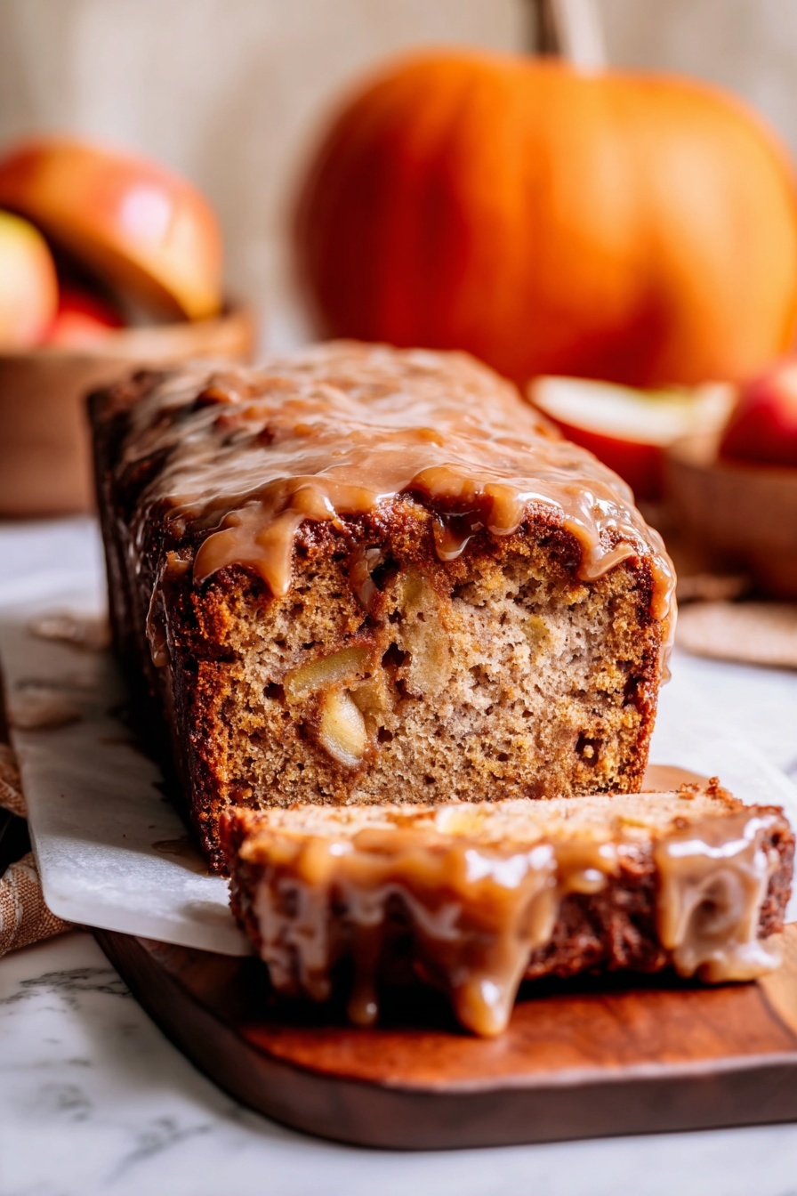A loaf cake sits on a wooden board over a white marbled surface, sliced at the front showing its dense, light brown inside with visible chunks of apple. The top layer is a thick, sticky, darker brown glaze that drips down the sides, giving the cake a shiny look. In the background, there is a small orange pumpkin and a wooden bowl with apples that add warm tones to the image. The photo is warm and cozy, focusing tightly on the textures of the cake and glaze, with a soft, light blur in the background. photo taken with an iphone --ar 2:3 --v 7 - Amish Apple Fritter Bread, apple fritter bread, warm apple dessert, cinnamon apple bread, comforting fall bread