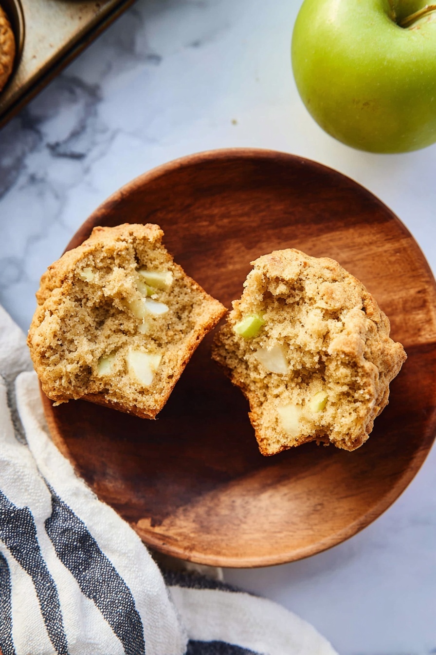 A round wooden plate on a white marbled surface holds a split muffin showing its soft, light brown crumb mixed with small white and slightly green apple pieces inside. To the right, a green apple sits partially in frame, and a striped white and dark blue cloth is partly visible beneath the plate and muffin tray corner. The muffin has a slightly golden crust with a rough texture. photo taken with an iphone --ar 2:3 --v 7 - Healthy Apple Muffins, healthy apple muffins recipe, wholesome apple muffins, nutritious apple muffin ideas,  apple muffin breakfast