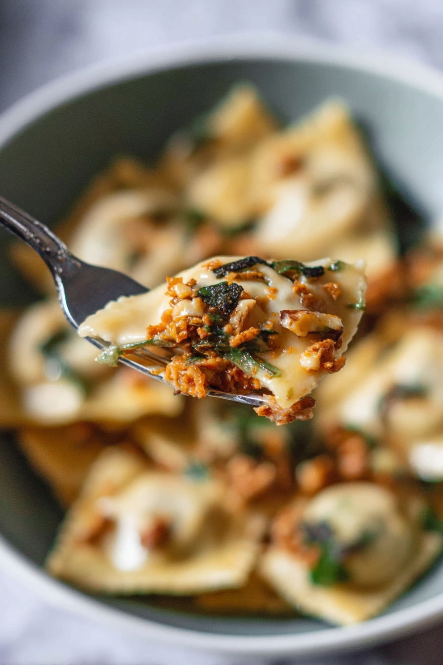 A close-up view shows a silver fork holding a piece of ravioli filled with a rich orange-brown meat mixture, topped with small dark green herb pieces and bits of light brown walnuts. In the blurred background, there is a white bowl filled with more ravioli, showing a mix of creamy beige pasta, white sauce, greens, and scattered walnut pieces, all set against a white marbled surface. The scene captures the soft texture of the ravioli and the fresh, rustic topping with a natural light that highlights the details. photo taken with an iphone --ar 2:3 --v 7 - Pumpkin Ravioli with Apple Sage Butter, pumpkin ravioli, homemade ravioli, fall pasta recipes, apple sage butter