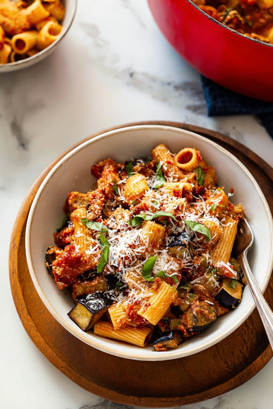 The image shows a white bowl placed on a round wooden tray on a white marbled surface. Inside the bowl, there are three main layers: the bottom layer has sautéed, golden-brown slices of eggplant, the middle layer consists of rigatoni pasta tubes mixed with a chunky red tomato sauce, and the top layer is sprinkled with shreds of white cheese and fresh green basil leaves. A silver fork is partially inserted into the pasta on the right side of the bowl. In the background and slightly to the left, a red pot filled with similar pasta can be seen. photo taken with an iphone --ar 2:3 --v 7 - Roasted Eggplant Pasta with Marinara, Eggplant pasta recipe, vegetarian pasta with roasted eggplant, easy eggplant pasta dish, healthy roasted eggplant recipe