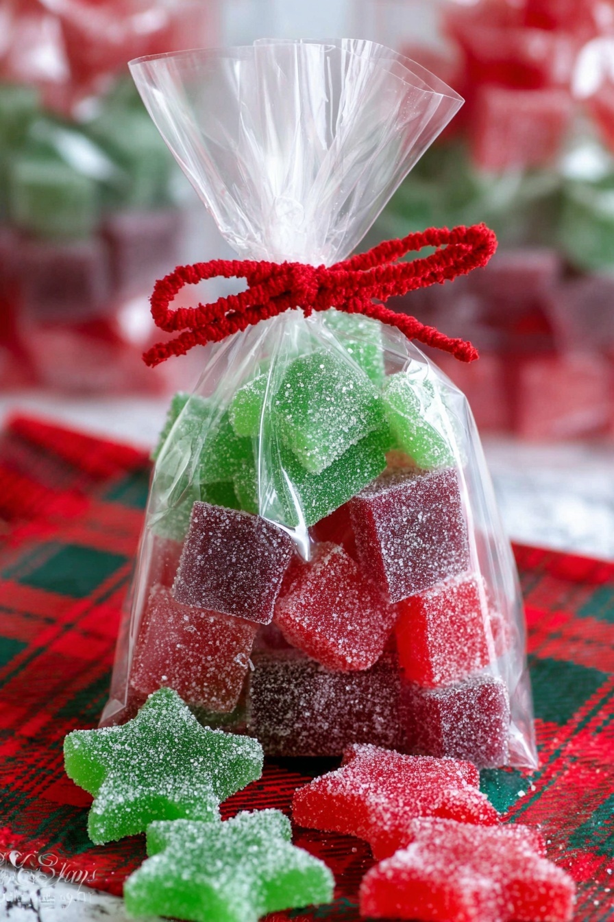 A clear plastic bag filled with stacked square sugar-coated gummies in red, green, and purple colors, arranged in alternating layers. There are also red and green star-shaped sugar-coated gummies at the bottom of the bag and scattered outside on a red and green plaid cloth. The bag is tied at the top with a red twist ribbon and decorated with a red felt scalloped band. The background shows a white marbled texture with blurred additional bags of similar candy. Photo taken with an iphone --ar 2:3 --v 7 - Homemade Gelatin Gumdrops, homemade gelatin gummies, chewy candy recipe, DIY gummy candies, customizable jello candies