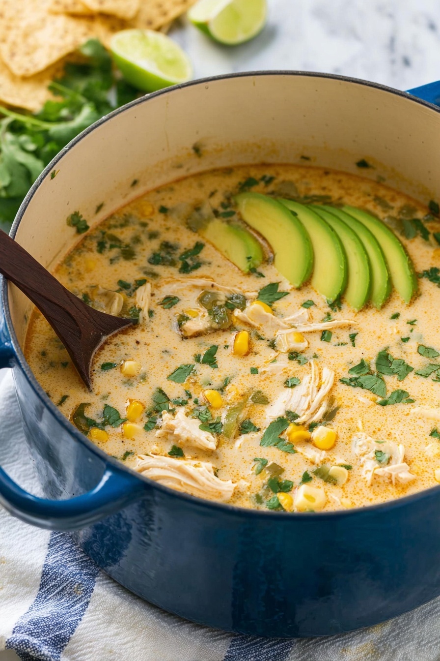A large blue pot filled with creamy soup that has a light orange color with green cilantro leaves scattered on top. Inside the soup, there are chunks of white chicken, pieces of yellow corn, and bits of green vegetables. On one side, thin slices of light green avocado are placed in a neat row, partially submerged in the soup. A dark wooden spoon is stirring the soup from the left side. The pot sits on a white cloth with blue stripes on a white marbled surface. In the background, there are some light beige tortilla chips and lime wedges. Photo taken with an iphone --ar 2:3 --v 7 - White Chicken Chili, White Chicken Chili recipe, easy white chicken chili, best white chicken chili, creamy chicken chili