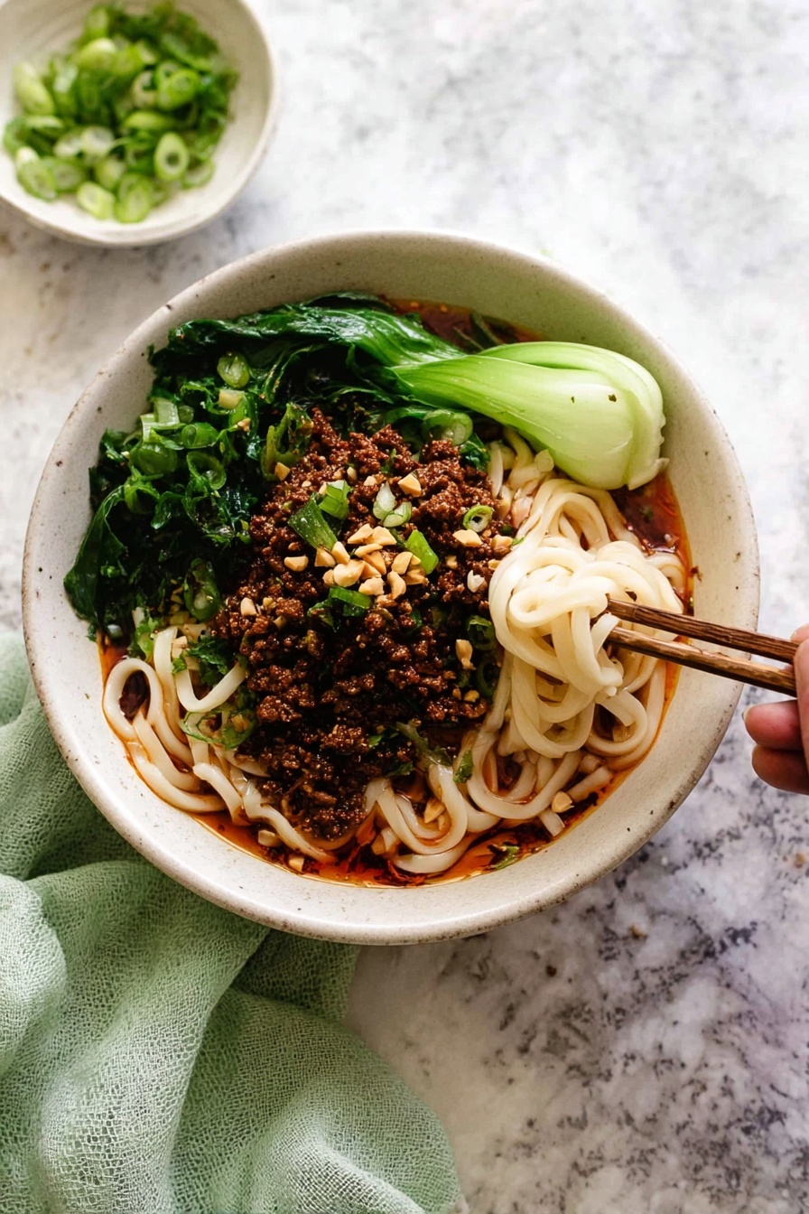 A white bowl filled with three main layers: at the bottom, thick white noodles mixed with a red oily sauce that gives a shiny texture; on top, a layer of dark brown minced meat scattered evenly; around the edges, bright green leafy vegetables adding fresh color; garnished with sliced light green scallions and small beige crushed nuts. A pair of dark brown chopsticks hold a small twisted bunch of noodles dipped in the sauce at the right side of the bowl. Some scallions and nut bits are scattered on the dark surface around the bowl. photo taken with an iphone --ar 2:3 --v 7 - Authentic Dan Dan Noodles with Pork and Sesame, Dan Dan Noodles recipe, spicy Sichuan noodles, Chinese pork noodle dish, sesame and chili noodle bowl
