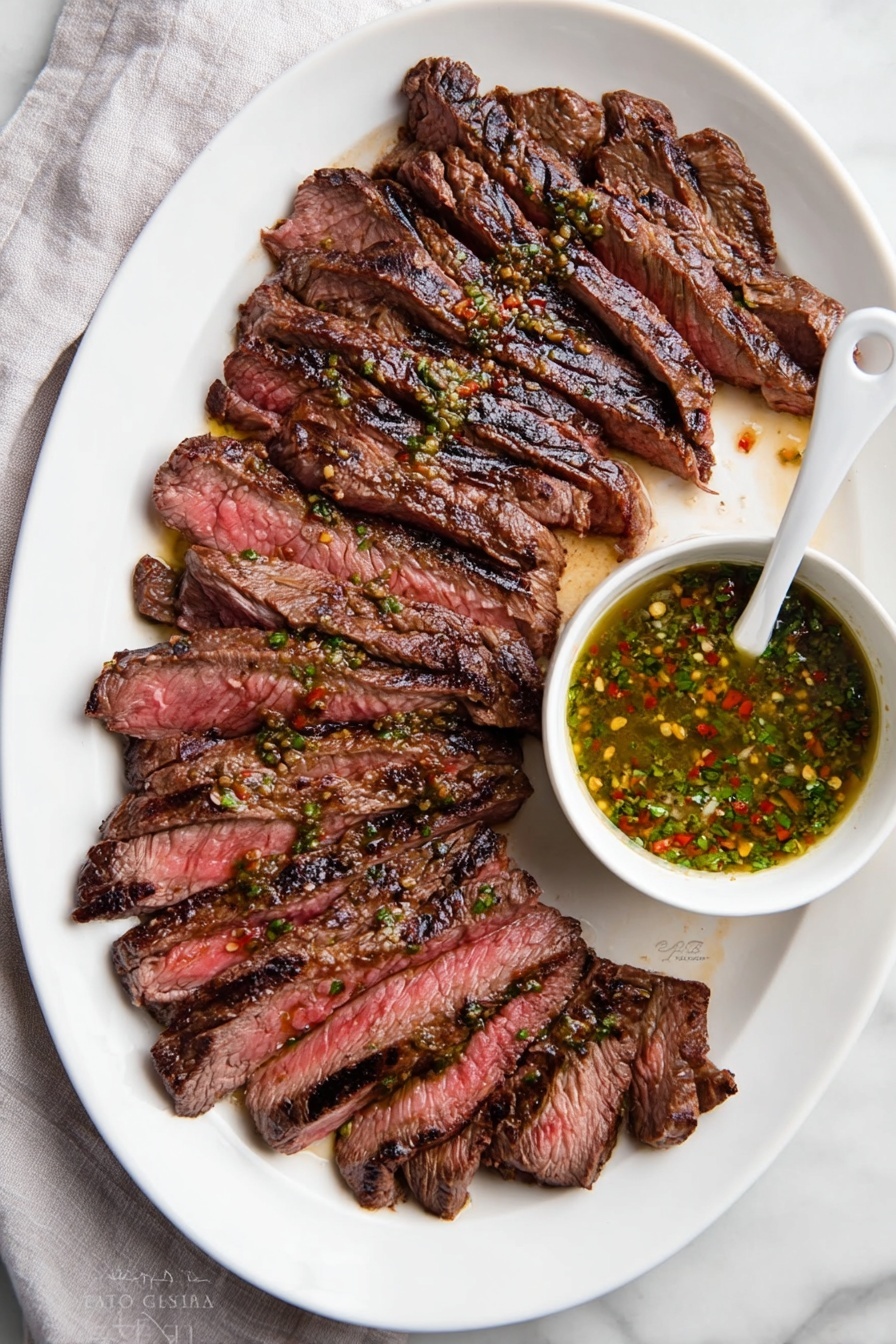 A white oval plate holds about twelve slices of grilled steak arranged in a slightly fanned out layer from left to right. The steak slices are medium-rare, showing a mix of deep brown outer edges with char marks and a pink-red center. To the right side of the plate, a small white bowl filled with a green sauce, flecked with red and yellow spices, sits partially on the plate with a white spoon inside it. The plate is set on a white marbled surface with a light gray cloth partially visible on the left side. Photo taken with an iphone --ar 2:3 --v 7 - Chimichurri Flank Steak, flank steak with chimichurri, easy chimichurri steak recipe, flavorful steak dinner, quick steak marinade