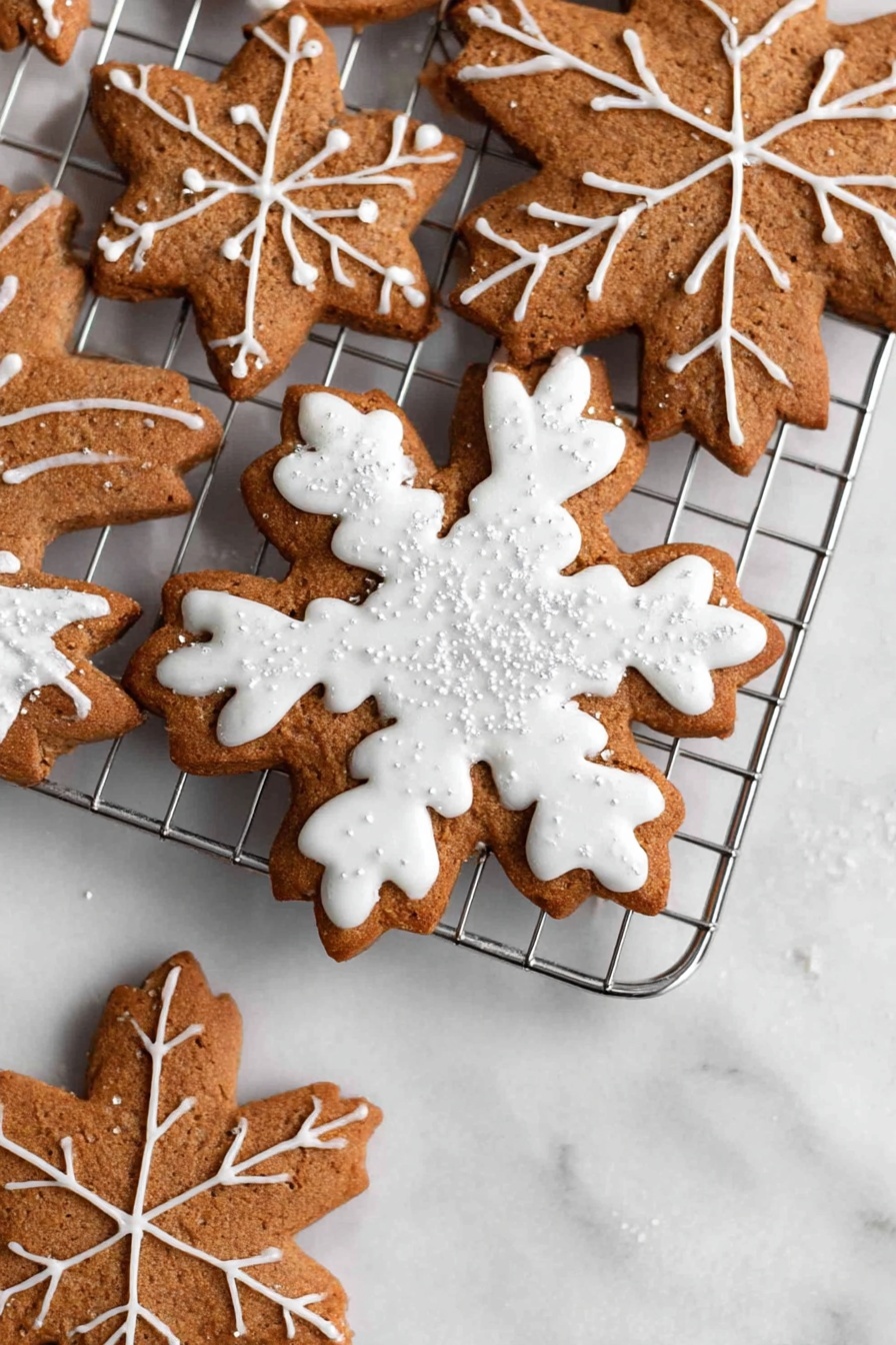 The image shows three snowflake-shaped cookies stacked on a silver cooling rack placed on a white marbled surface. The cookies are brown with detailed white icing decoration. The top cookie has thick white icing covering the shape with a leaf vein pattern on each arm and small shiny sugar crystals scattered on top. The two cookies underneath have thinner white icing lines creating a delicate snowflake design with crisscross patterns. The cookies’ texture looks soft yet firm with visible edges. Photo taken with an iphone --ar 2:3 --v 7 - Vegan Gingerbread Cookies, plant-based gingerbread cookies, healthy holiday cookies, vegan holiday treats, gluten-free gingerbread cookies