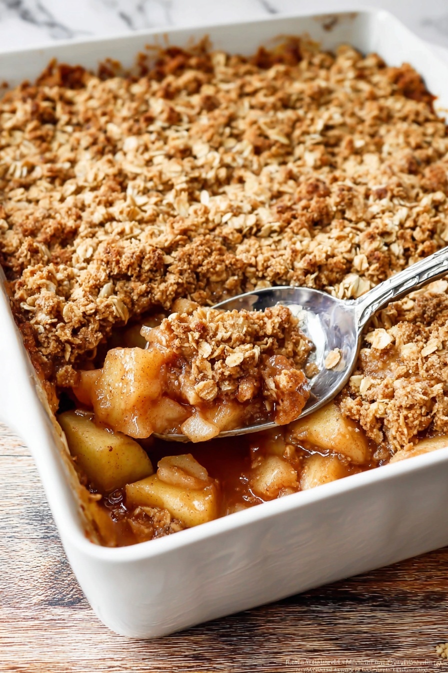 The image shows a white baking dish filled with a baked apple dessert. The bottom layer consists of soft, cooked apple chunks in a golden-brown syrup. Above this is a thick layer of crumbly oat topping, golden and slightly toasted with visible oats and brown sugar. A silver spoon scoops out some of the dessert from the bottom right corner, revealing the juicy apple filling beneath the crunchy topping. The white dish is set on a wooden surface with a white marbled texture in the background. Photo taken with an iphone --ar 2:3 --v 7 - Ultimate Apple Crisp, apple crisp dessert, cozy apple dessert, classic apple crisp recipe, easy apple crumble