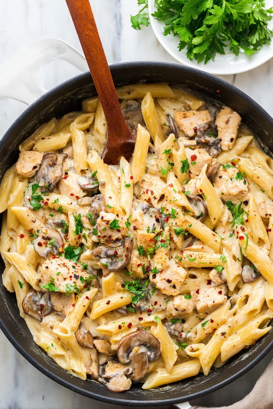 A black pan filled with creamy pasta made of three main layers: the base layer of pale yellow penne pasta, the middle layer of light brown cooked mushroom slices, and the top layer of small light beige pieces of chicken mixed in a creamy sauce. Green parsley leaves are scattered on top, adding bright green spots, with tiny black and red pepper flakes sprinkled over all. A wooden spoon rests inside the pan, partially covered by the pasta. The pan is placed on a white marbled surface, with a small white plate of fresh parsley leaves in the top corner. photo taken with an iphone --ar 2:3 --v 7 - Creamy Chicken Marsala Pasta, Chicken Marsala Pasta, creamy chicken pasta, easy chicken mushroom pasta, quick dinner recipes