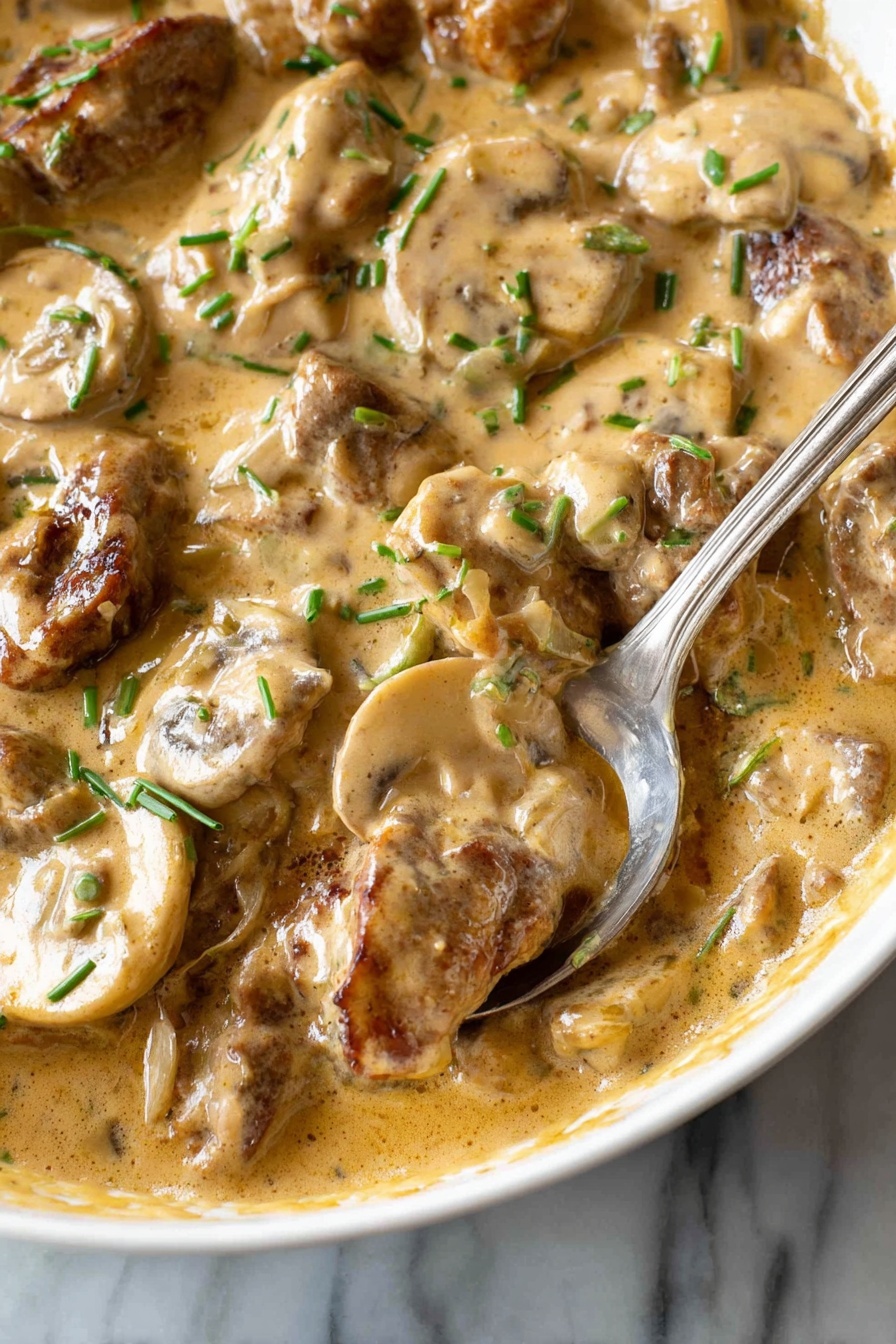 This image shows a close-up of a creamy dish with several layers visible. The base layer is a thick, light brown sauce with a smooth, slightly shiny texture, pooling around the other ingredients. Mixed in the sauce are slices of light beige mushrooms with soft, rounded edges and a slightly wet look. Also present are pieces of browned meat, which have a slightly rough texture and are coated in the same sauce. Small green chive pieces are scattered on top, adding little spots of bright green color throughout. A silver spoon is partially dipped into the dish, resting on the edge of a white bowl. The background is a white marbled surface. Photo taken with an iphone --ar 2:3 --v 7 - Quick Beef Stroganoff with Mushrooms, easy beef stroganoff, creamy mushroom beef dinner, fast weeknight beef recipes, hearty beef mushroom dish