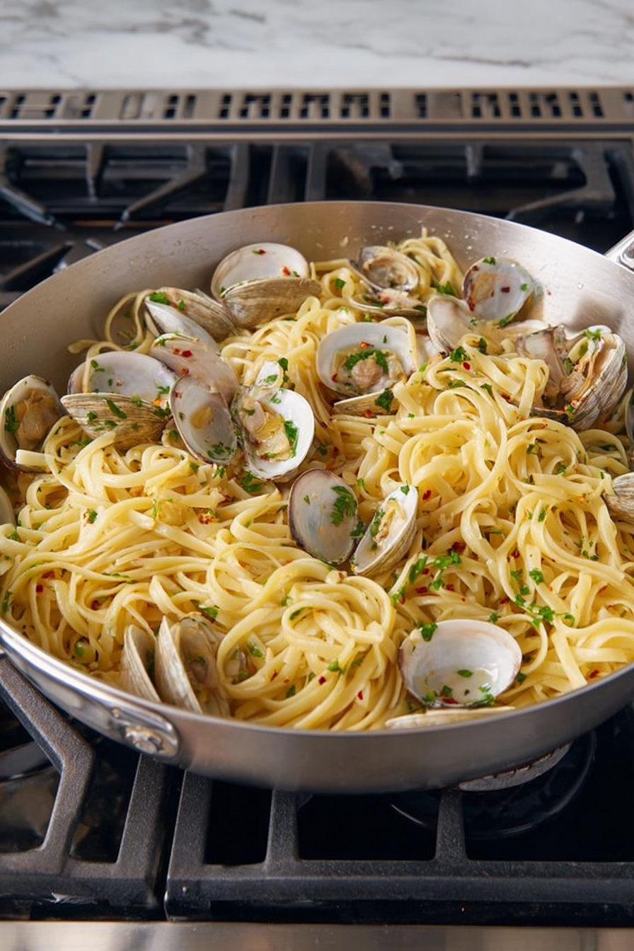 A large silver pan filled with cooked linguine pasta mixed with open clams distributed evenly throughout. The pasta is light yellow, soft and slightly shiny from sauce, with chopped green herbs sprinkled on top for color and freshness. Small bits of garlic and red chili flakes are visible mixed into the pasta, adding texture and flavor. The pan sits on a black stove with metal grates, surrounded by a white marbled surface. photo taken with an iphone --ar 2:3 --v 7 - Linguine with Clams and Lemon, clam linguine recipe, lemon pasta with clams, seafood pasta recipes, easy clam linguine