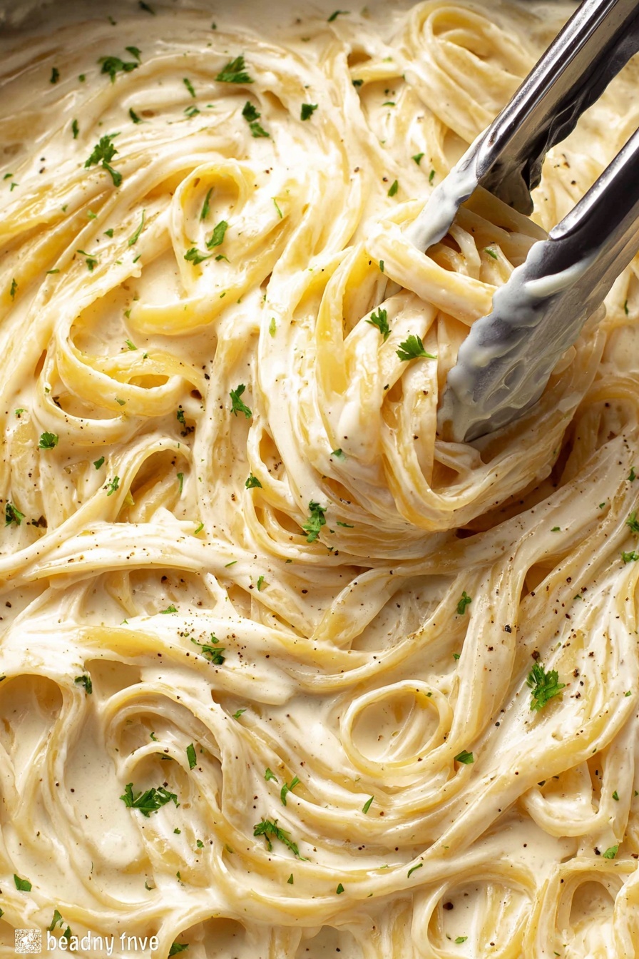 A close-up view of creamy fettuccine pasta in a thick white sauce filling the whole frame, with the pasta strands twisted and intertwined. The sauce is smooth and glossy with a light beige color and flecks of black pepper and small green parsley pieces sprinkled on top. Silver tongs are grabbing a bunch of pasta near the center, showing the rich texture of the sauce coating the noodles. The background is a white marbled texture. photo taken with an iphone --ar 2:3 --v 7 - Creamy Chicken Alfredo Pasta, chicken Alfredo pasta recipe, homemade Alfredo sauce, easy chicken pasta, comforting creamy pasta dish