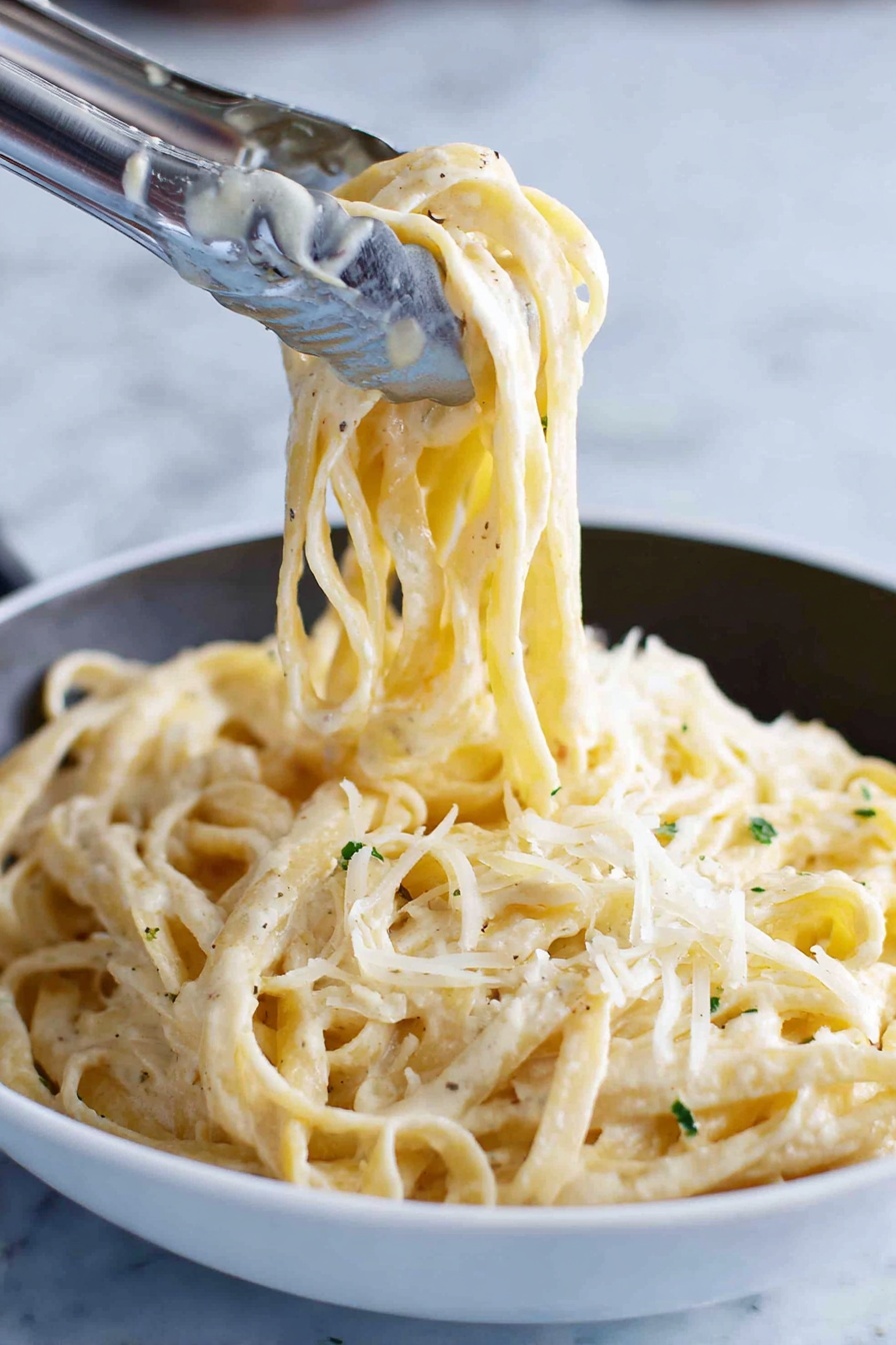 A close-up image shows thick noodles coated with a creamy white sauce, twirled and lifted by silver tongs above a white bowl filled with the same noodles. The noodles have a slightly glossy texture from the sauce and small green herb bits sprinkled throughout. Light shreds of pale cheese are scattered on top, adding a soft, fine texture contrast against the smooth noodles. The background is a white marbled surface. photo taken with an iphone --ar 2:3 --v 7 - Creamy Fettuccine Alfredo, homemade Alfredo sauce, easy pasta dinner, creamy pasta recipes, indulgent Italian pasta