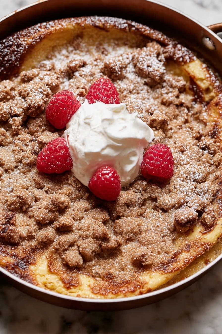A round dessert with two main layers is shown in a pan. The bottom layer is a golden-brown, slightly risen cake with some darker browned spots. On top is a crumbly, light brown streusel layer that covers most of the surface unevenly. There is a dollop of white cream in the center with four bright red raspberries placed around and on it. A light dusting of powdered sugar is scattered over parts of the streusel and cake edges. The pan sits on a white marbled surface. photo taken with an iphone --ar 2:3 --v 7 - Brioche French Toast Casserole, easiest French toast casserole, holiday brunch recipes, make-ahead breakfast ideas, decadent breakfast casserole