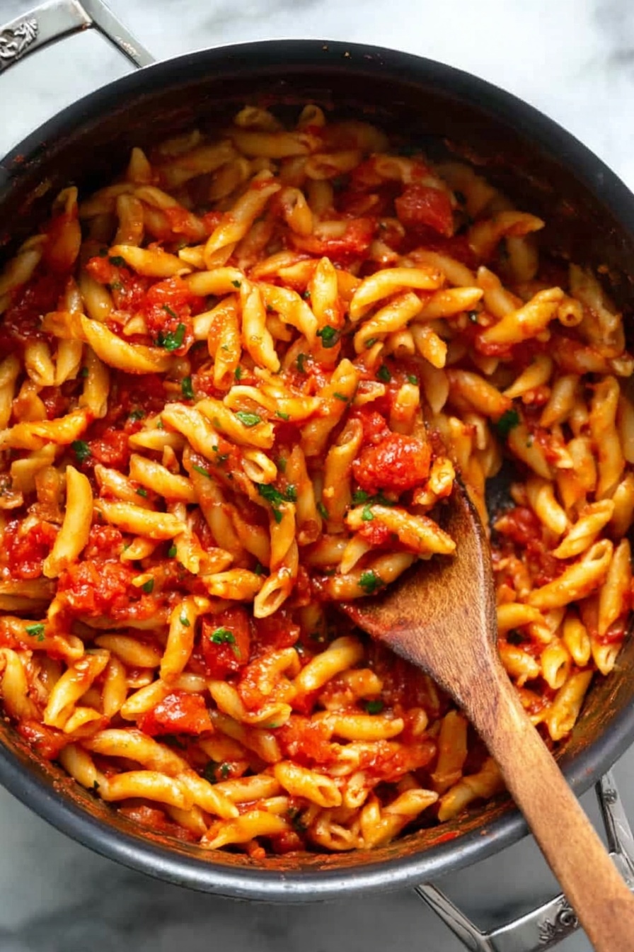 A black pot filled with a single layer of short, twisted pasta all covered evenly in a bright red tomato sauce with small bits of tomatoes and green herbs mixed in. A woman’s wooden spoon is stuck deep in the middle, lifting some pasta, showing the sauce clinging to the noodles. The pot is on a white marbled surface, the pasta looks soft and shiny with sauce. Photo taken with an iphone --ar 2:3 --v 7 - Spicy Penne Arrabbiata, spicy pasta, easy Italian pasta, quick dinner recipes, flavorful penne pasta