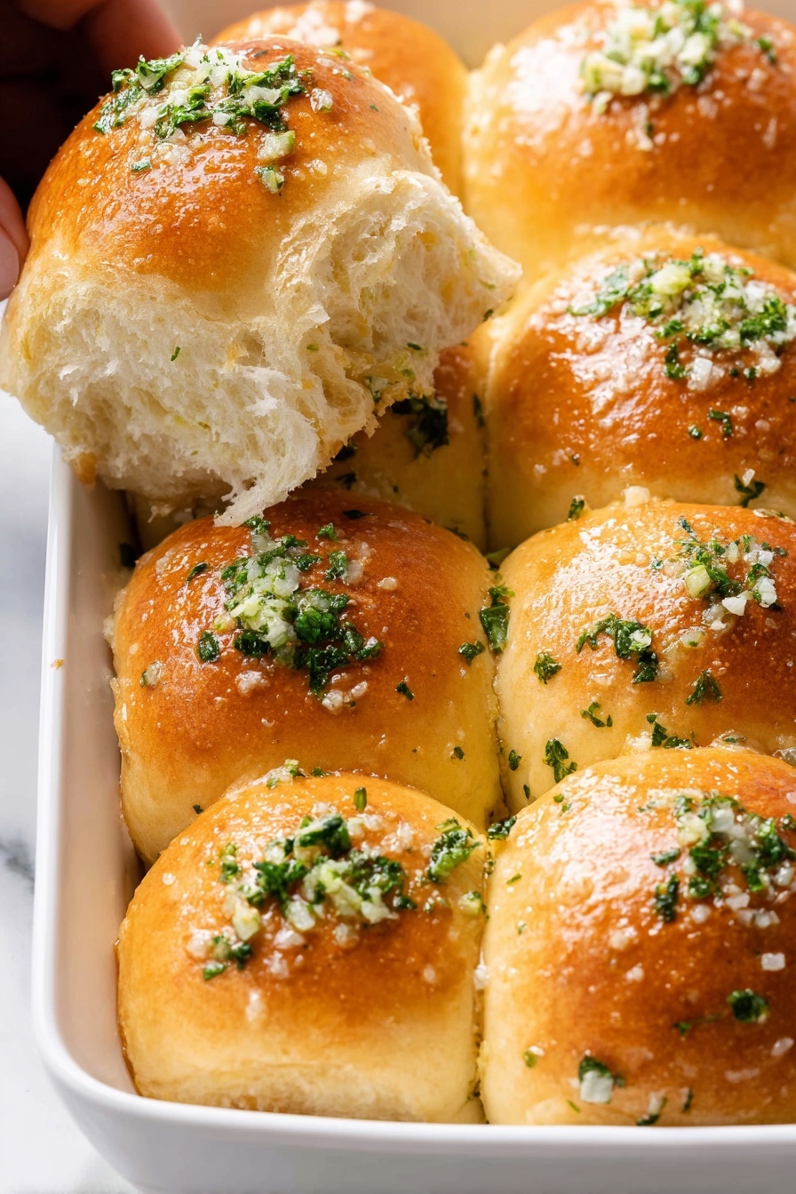 The image shows a white dish filled with seven shiny, golden-brown dinner rolls bunched together in two rows. Each soft roll is topped with small bits of green parsley and finely chopped garlic, sprinkled evenly with coarse salt. One roll is slightly pulled away, revealing a fluffy, light inside with a soft texture. There is a woman's hand partially holding this separated roll on the top left of the frame. The background and surface are a white marbled texture. photo taken with an iphone --ar 2:3 --v 7 - Garlic Bread Rolls Thanksgiving, garlic bread rolls, easy holiday bread, buttery garlic rolls, Thanksgiving side dishes