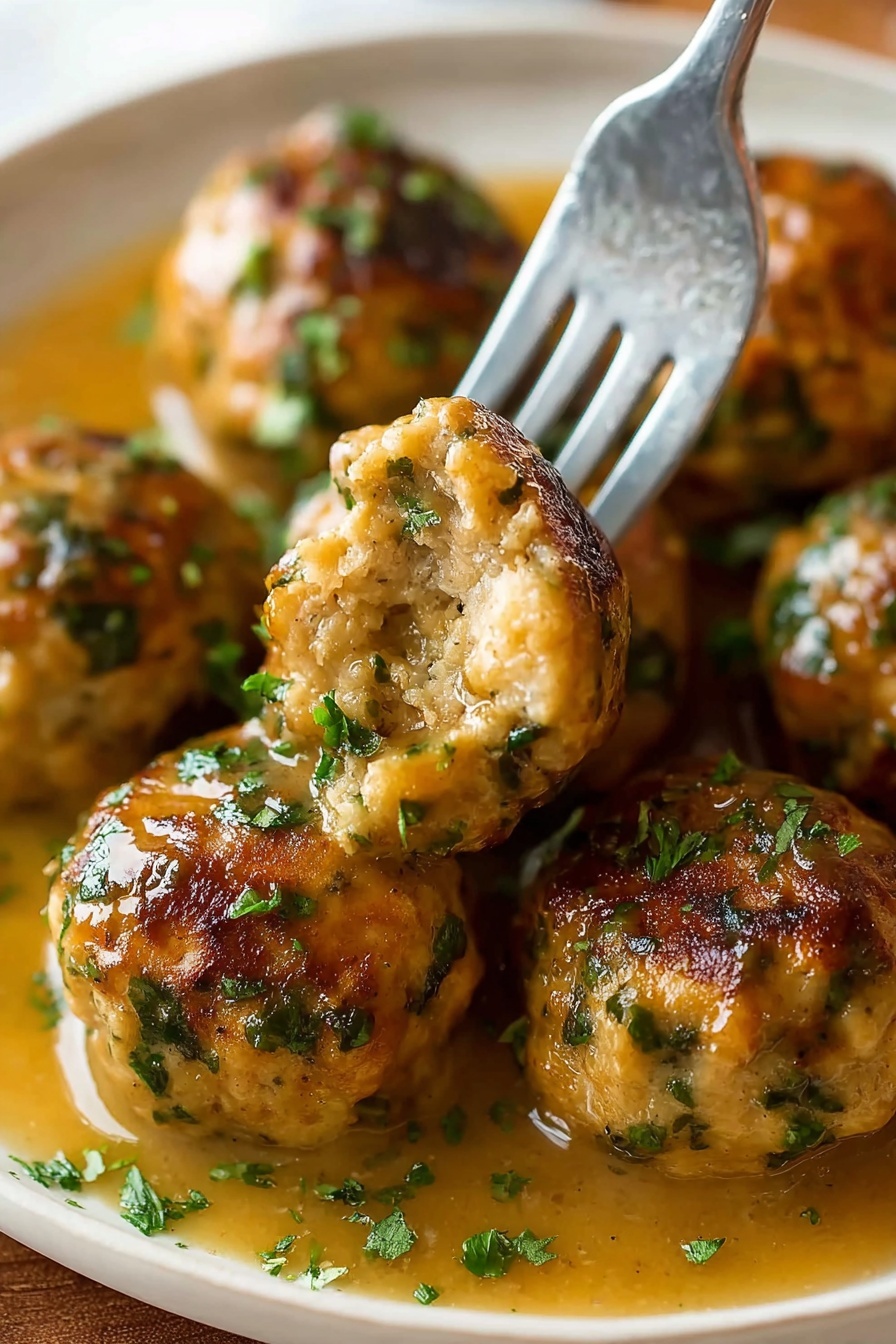 The image shows several round meatballs with a golden-brown crust and green herb bits scattered through them, resting in a light brown gravy. A silver fork holds one meatball with a bite taken out, showing a soft and moist inside with visible herbs. The meatballs are arranged closely on a white plate with some chopped green herbs sprinkled on top and the gravy pooling around them. The background is a white marbled texture photo taken with an iphone --ar 2:3 --v 7 - Stuffing Balls, Holiday Side Dishes, Easy Stuffing Balls, Crispy Stuffing Balls, Thanksgiving Side Dishes