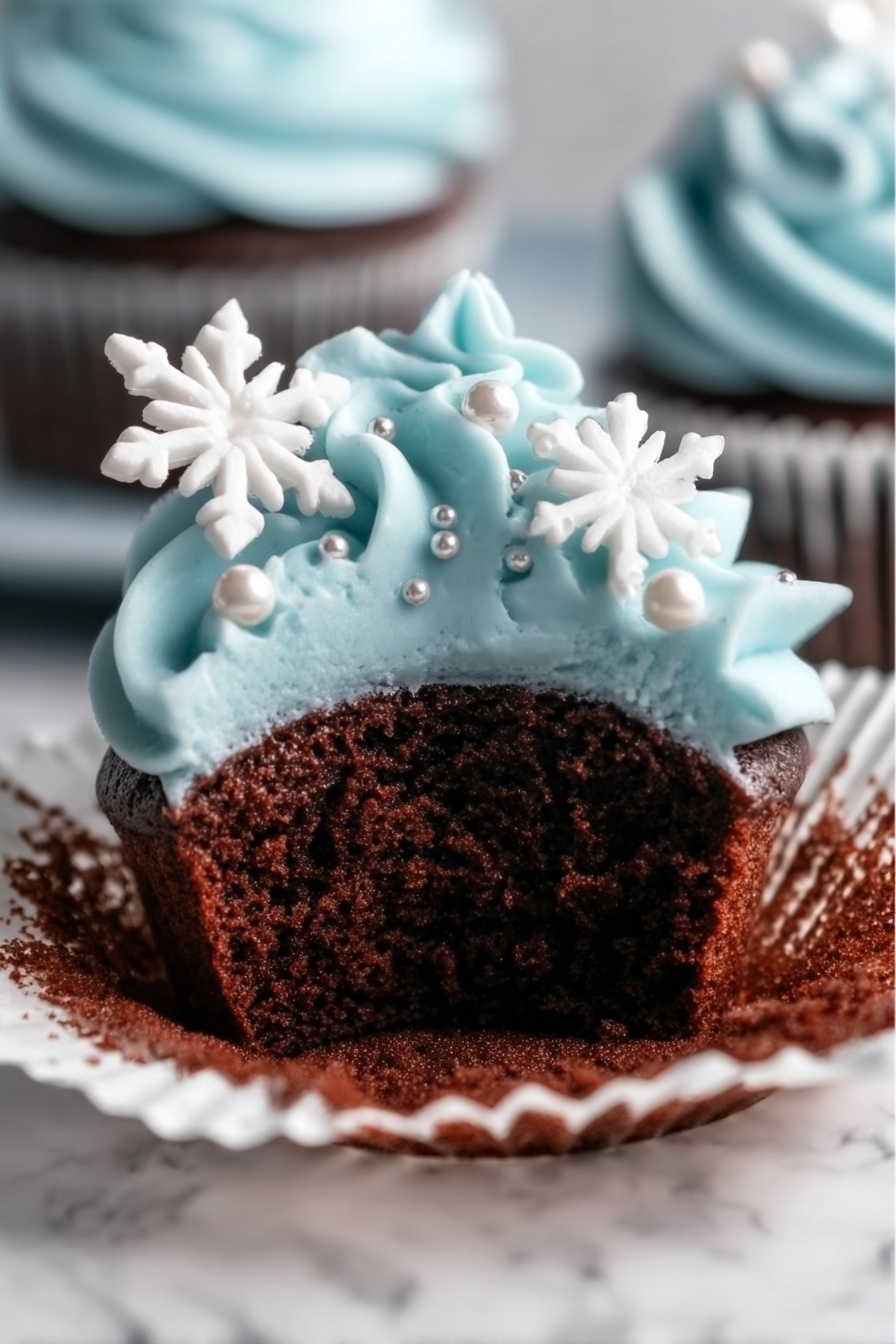 A close-up view of a chocolate cupcake with a white cupcake liner partially peeled back, showing the soft, dark brown cake inside. The top layer is covered with smooth, light blue frosting piped in swirls, decorated with small silver pearls and two white snowflake-shaped sprinkles placed on the side. The cupcake is placed on a white marbled surface. Photo taken with an iphone --ar 2:3 --v 7 - Winter Wonderland Chocolate Cupcakes, chocolate cupcakes with snowflake buttercream, winter themed chocolate cupcake recipe, festive chocolate cupcake ideas, cozy winter dessert recipes
