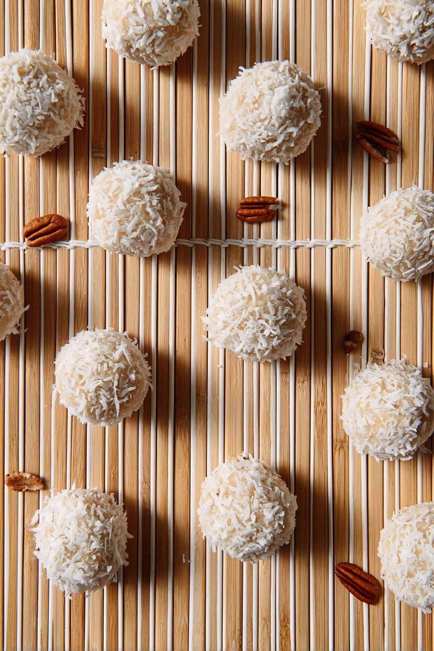 The image shows multiple round balls covered with white coconut flakes arranged on a light bamboo matt with parallel white strings running vertically. The balls have a beige base visible through the coconut flakes and are scattered evenly across the frame. There are small pecan nuts placed randomly around the balls, adding a brown color contrast to the scene. The overall look is clean with a natural, textured feel. Photo taken with an iphone --ar 2:3 --v 7 - Orange Coconut Snowball Cookies, tropical cookie recipes, coconut ball cookies, easy holiday cookies, citrus dessert recipes
