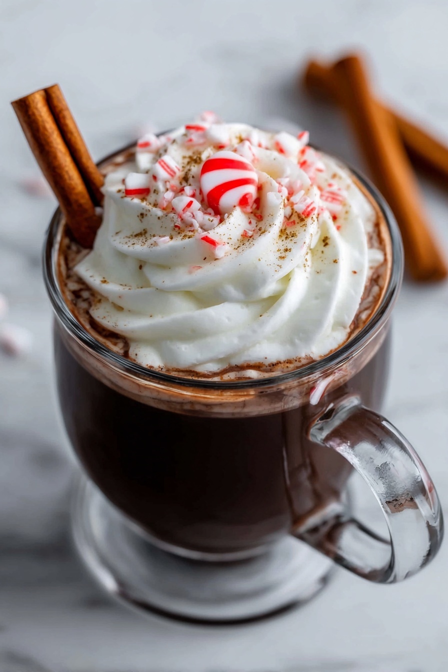 A clear glass mug filled with dark hot chocolate topped with a large swirl of white whipped cream. On top of the cream is a small red and white peppermint candy in the center, surrounded by some small pieces of crushed candy cane and fine brown sprinkles. A brown cinnamon stick is resting against the side inside the mug. The mug is placed on a white marbled surface. In the background, two more cinnamon sticks lie on the surface out of focus. A woman's hand is holding the handle of the mug. photo taken with an iphone --ar 2:3 --v 7 - Festive Peppermint Hot Chocolate, Peppermint Hot Chocolate, Holiday Hot Chocolate, Winter Warmers, Christmas Hot Chocolate
