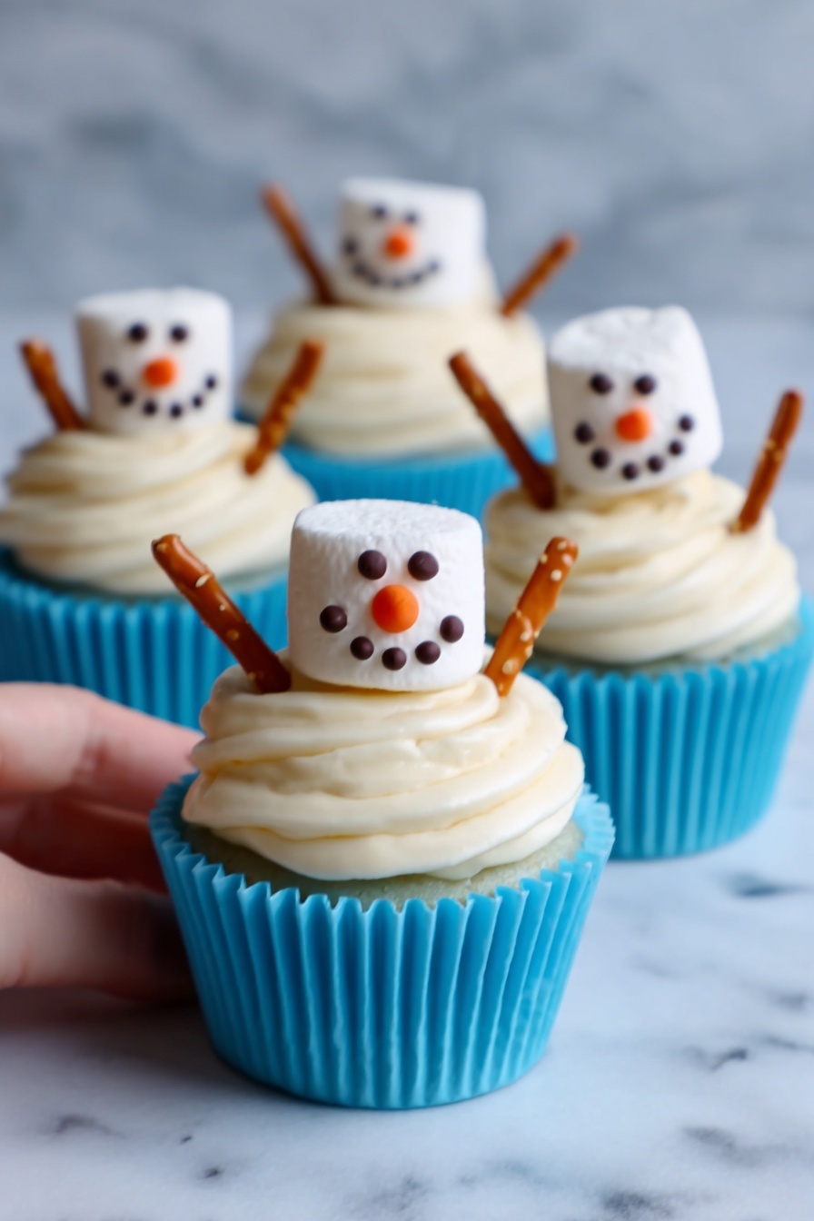 The image shows three cupcakes in white cupcake liners with bright blue bases, each topped with thick, creamy white frosting swirled in layers. On top of each cupcake sits a large marshmallow decorated like a snowman face with small dark chocolate or icing dots for eyes and mouth, and an orange triangular nose. Two pretzel sticks are inserted on opposite sides of the frosting, looking like the snowman's arms. The cupcakes are placed on a white marbled surface, with one woman's hand gently holding the front cupcake. The scene has bright, soft lighting. photo taken with an iphone --ar 2:3 --v 7 - Snowman Cupcake Muffins, festive holiday cupcakes, Christmas cupcake ideas, winter baking treats, adorable holiday muffins