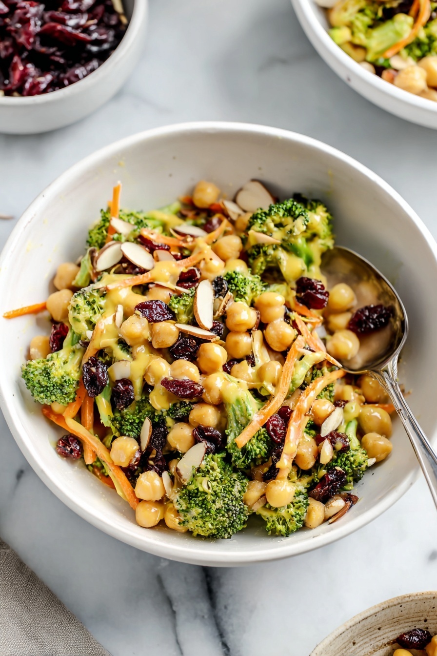 A white bowl filled with a colorful salad that has three main layers: the base layer is light beige chickpeas scattered throughout, the middle layer shows small bright green broccoli florets and chopped stems, and the top layer consists of thin orange carrot strips, dark red dried cranberries, and light brown almond slices evenly spread. A creamy yellow dressing lightly coats the salad, giving a soft shine to the vegetables and chickpeas. A silver spoon is placed inside the bowl resting among the salad. The bowl sits on a white marbled surface, and in the background, parts of another white bowl and a white cup filled with dark red dried cranberries are visible. Photo taken with an iphone --ar 2:3 --v 7 - Vegan Curried Broccoli Chickpea Salad, vegan salad recipes, healthy chickpea salad, plant-based broccoli salad, quick vegan lunch ideas
