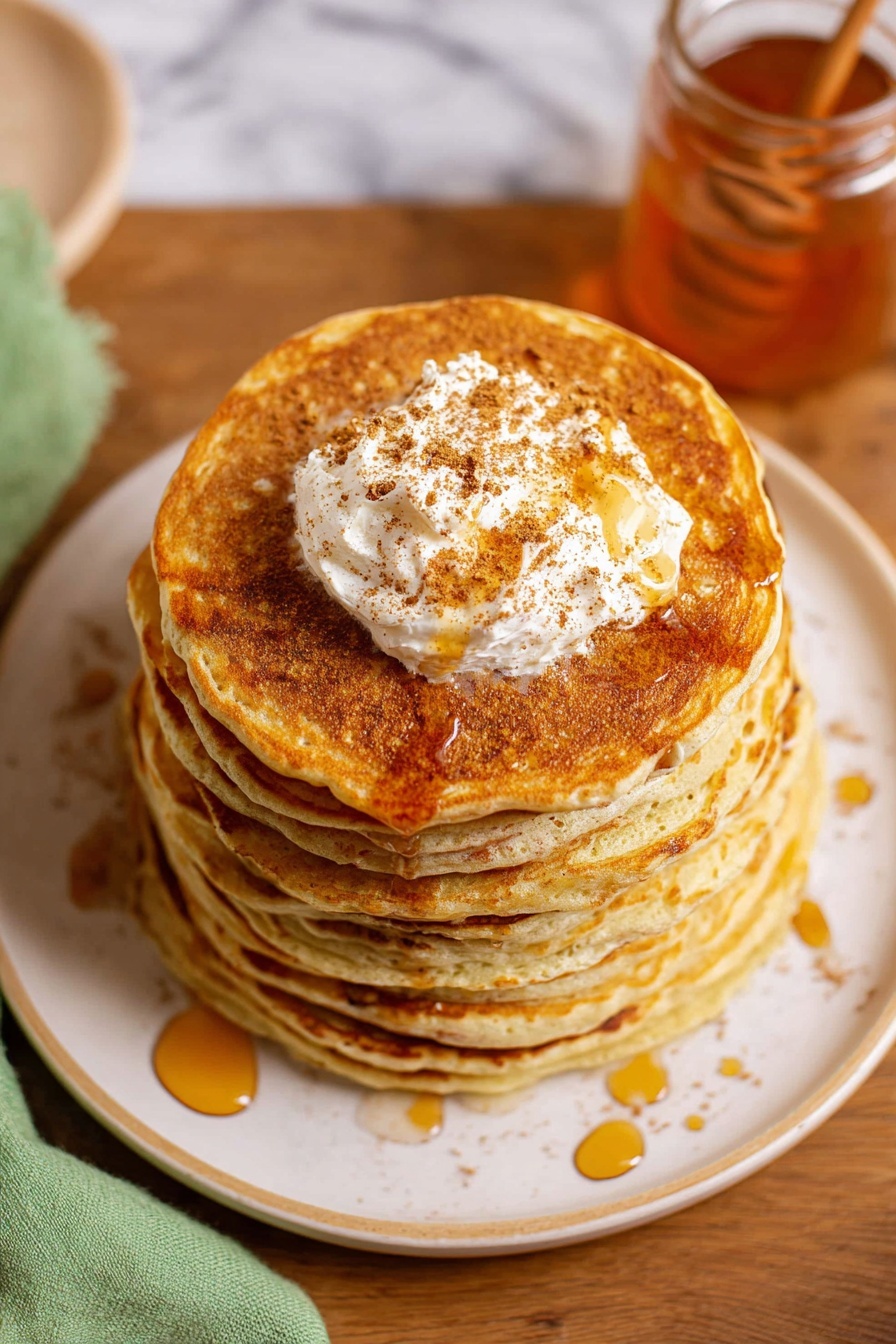 A stack of seven golden brown pancakes sits centered on a white plate with a subtle rim design, each pancake thick and fluffy with a smooth texture. On top of the stack is a generous dollop of cream-colored butter sprinkled with brown cinnamon powder, melting slightly with amber syrup drizzled over the pancakes in thin, uneven lines. The plate rests on a wooden table with a translucent glass jar of honey nearby, and a light green cloth is partially visible on the left side. The background features a white marbled texture. photo taken with an iphone --ar 2:3 --v 7 - Creamy Cinnamon Butter Spread, cinnamon butter spread, cinnamon butter recipe, flavored butter, breakfast spread