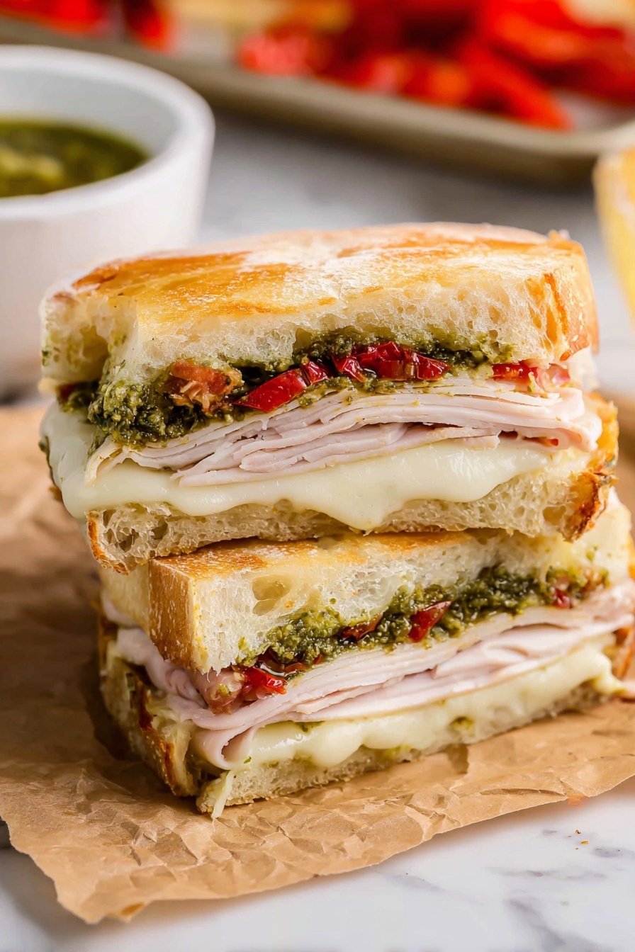 The image shows a sandwich cut in half and stacked, placed on crinkled brown paper against a white marbled surface. The sandwich has three main layers of fillings. The bottom layer consists of melted white cheese with a soft texture. Above it is a layer of thin, light pink slices of turkey. On top of the turkey, there is a green layer of pesto sauce mixed with small pieces of red roasted peppers. The sandwich is held together by two thick slices of golden brown, slightly crusty bread with a soft inside, forming the top and bottom of the sandwich. In the background, there is a white bowl with green sauce and part of a baking tray with more red roasted peppers visible. Photo taken with an iphone --ar 2:3 --v 7 - Copycat Starbucks Turkey Pesto Sandwich, Starbucks inspired turkey pesto sandwich, homemade turkey pesto sandwich, easy turkey pesto sandwich, best turkey pesto sandwich recipe